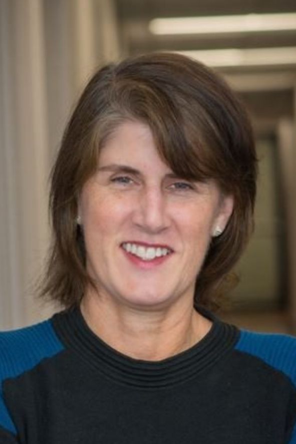A portrait of a smiling middle-aged woman with shoulder-length brown hair, wearing a black and blue top. She stands indoors with a soft-focus background.