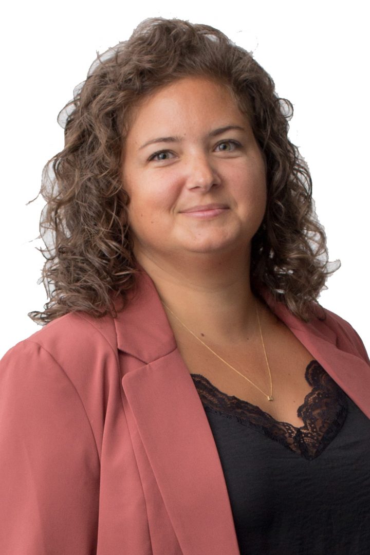 A woman with curly hair wearing a pink blazer over a black top with lace detail, a delicate necklace, and a subtle smile, against a white background.
