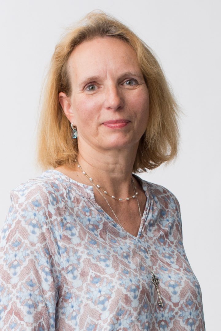 A headshot of a middle-aged woman with blond hair and blue floral blouse, wearing earrings, a necklace, and a pendant, on a white background.