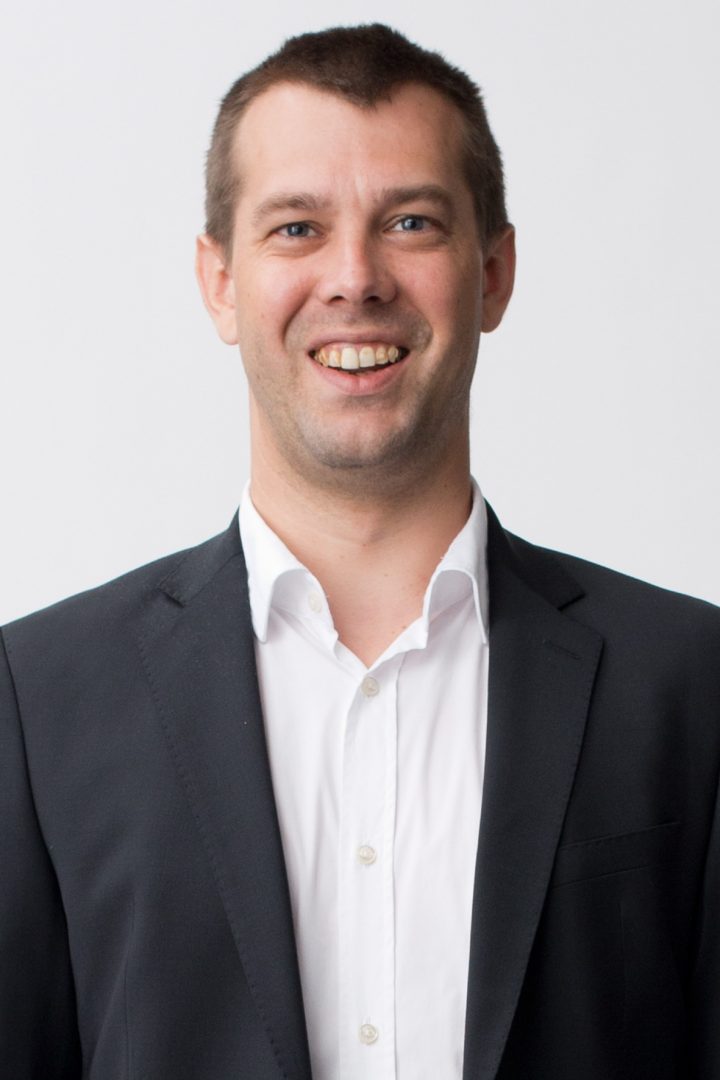 A smiling man with short hair wearing a dark blazer and white shirt against a white background.