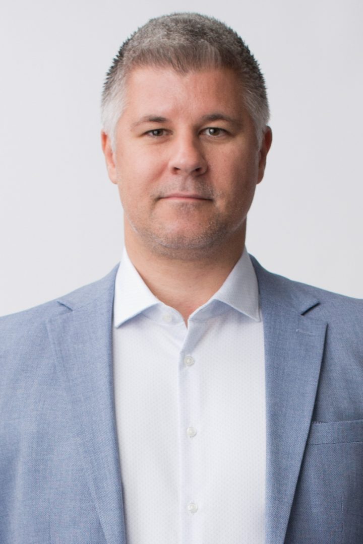 A professional headshot of a middle-aged man with short grey hair, wearing a light blue blazer over a white dress shirt, against a white background.