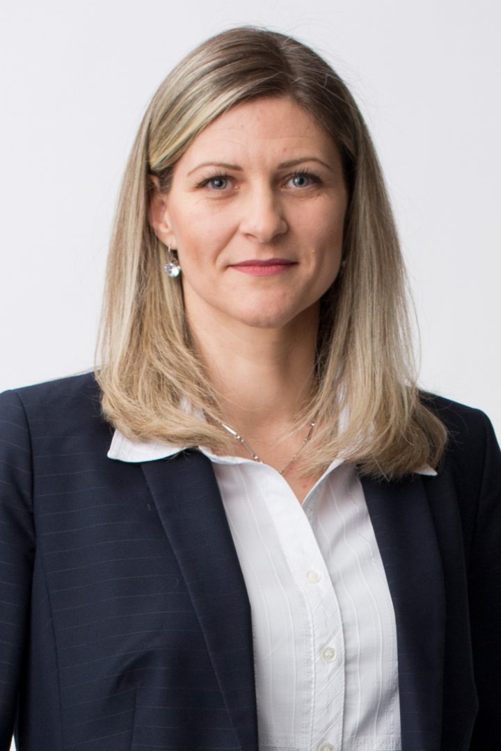 A professional headshot of a woman with shoulder-length blond hair, wearing a navy blue suit jacket, a white blouse, and pearl earrings against a white background.