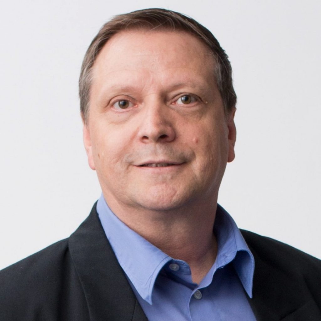 A middle-aged man with a slight smile, wearing a black blazer over a blue button-up shirt, against a white background.