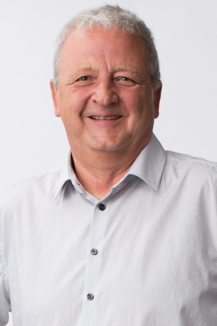 A smiling older man with gray hair, wearing a buttoned light gray shirt against a white background.