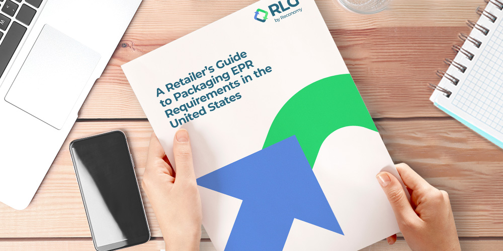 Hands holding a booklet titled "A Retailer's Guide to Packaging EPR Requirements in the United States" on a wooden desk beside a laptop, smartphone, and spiral-bound notebook.