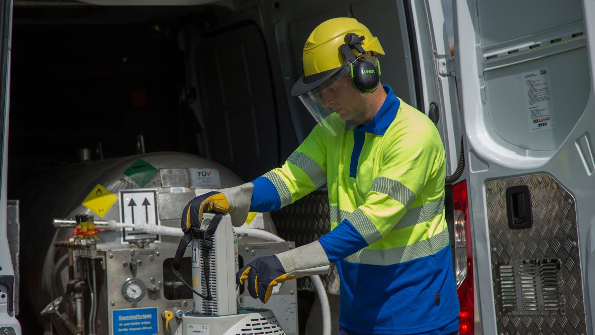 A worker wearing a yellow hard hat, safety glasses, and high-visibility jacket operates equipment near an open van.