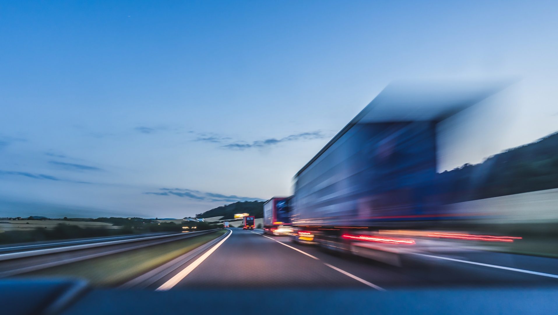 Motion blurred image of a highway with a large truck in the right lane and other vehicles with visible light trails captured during twilight.