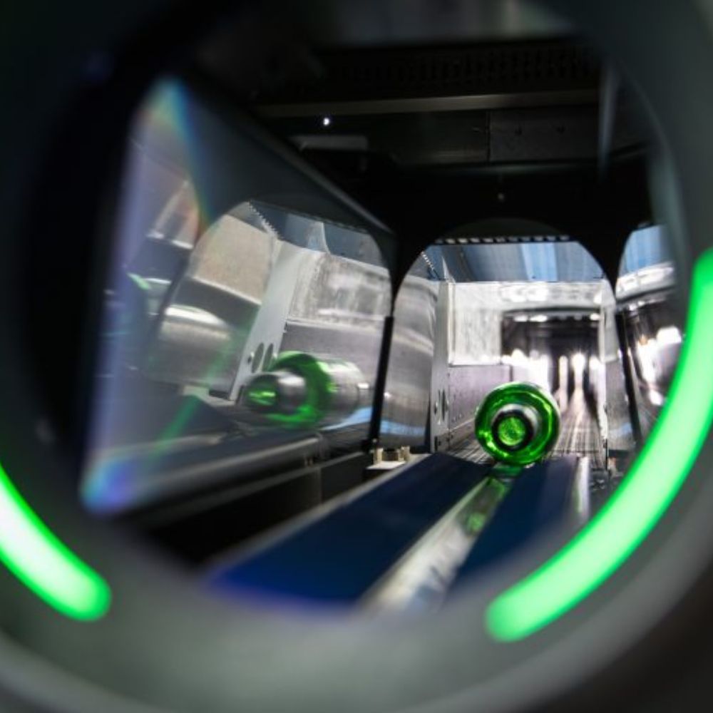 View down a production line showing a green glass bottle being conveyed through industrial machinery with reflective metal surfaces and green LED lights.