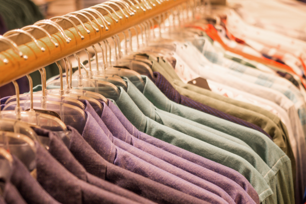 Row of men's collared shirts in purple, green and plaid patterns hanging closely on clear plastic hangers along a wooden clothing rack in a retail store