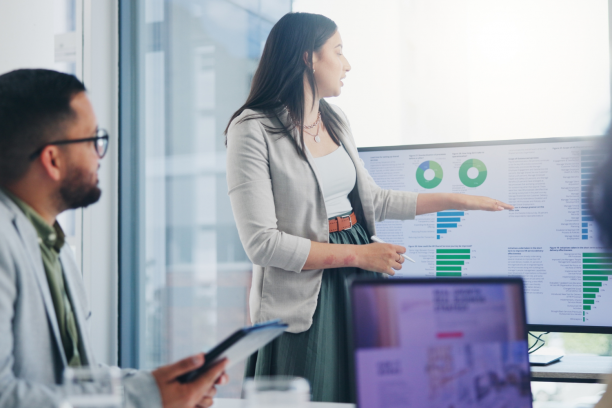 Woman in a gray blazer presenting in a glass-walled office, pointing at a monitor showing green pie and bar charts while a seated colleague with glasses holds a tablet and a laptop screen blurs in the foreground.